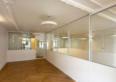Empty office corridor with glass-walled cubicles, white walls, and ornate ceiling moldings.